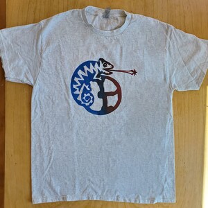 K&ouml;nnte beinhalten: Ein graues T-Shirt mit einem blauen und roten Cham&auml;leon-Motiv. Das Cham&auml;leon ist um einen Kreis mit einem Stern am Ende seiner Zunge gewickelt.