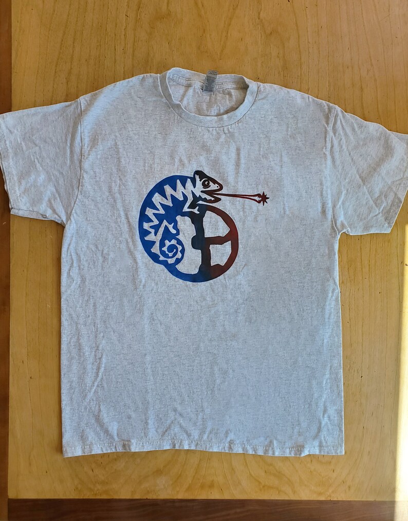 K&ouml;nnte beinhalten: Ein graues T-Shirt mit einem blauen und roten Cham&auml;leon-Motiv. Das Cham&auml;leon ist um einen Kreis mit einem Stern am Ende seiner Zunge gewickelt.