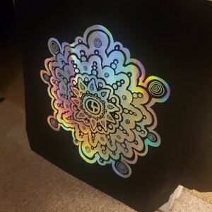 May include: A black tote bag with a large iridescent mandala design. The mandala is outlined in black and features a peace sign in the center.