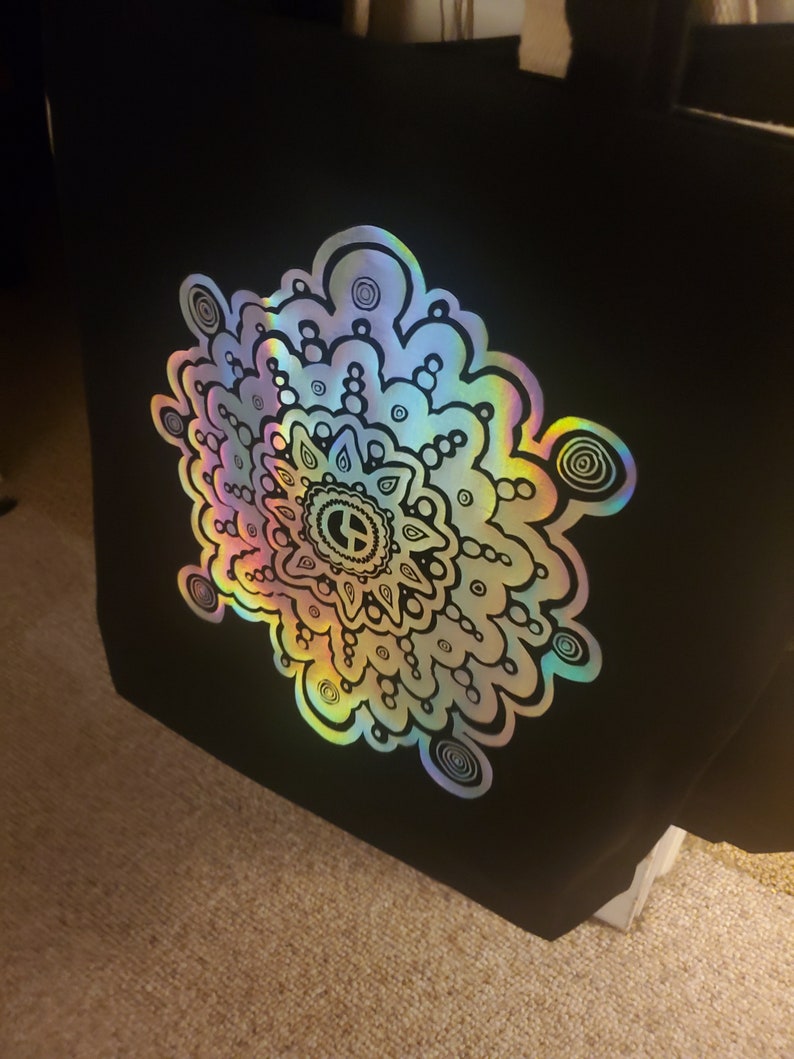 May include: A black tote bag with a large iridescent mandala design. The mandala is outlined in black and features a peace sign in the center.