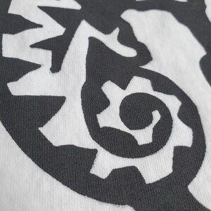 K&ouml;nnte beinhalten: Schwarz-wei&szlig;es Grafikdesign auf einem wei&szlig;en T-Shirt. Das Design zeigt eine stilisierte, abstrakte, zahnradf&ouml;rmige Form.
