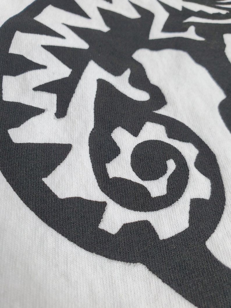 K&ouml;nnte beinhalten: Schwarz-wei&szlig;es Grafikdesign auf einem wei&szlig;en T-Shirt. Das Design zeigt eine stilisierte, abstrakte, zahnradf&ouml;rmige Form.