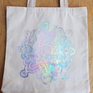 May include: A white canvas tote bag with a holographic iridescent mandala design. The mandala is a circular pattern with intricate details and a rainbow of colors.