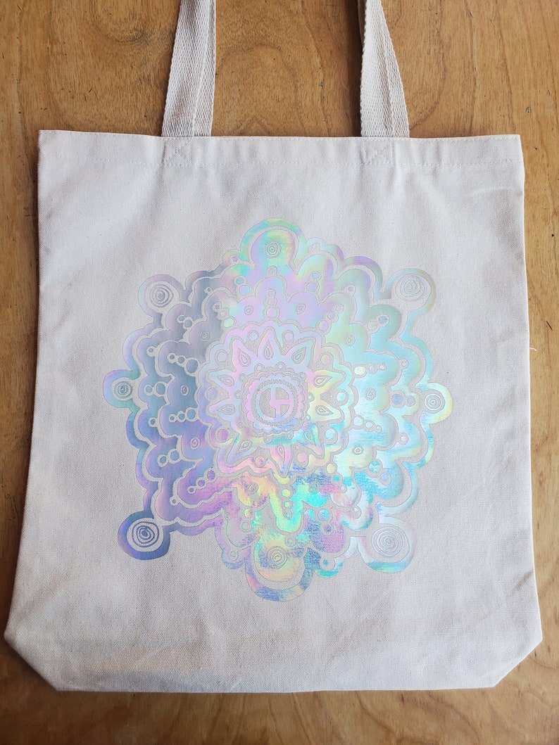 May include: A white canvas tote bag with a holographic iridescent mandala design. The mandala is a circular pattern with intricate details and a rainbow of colors.