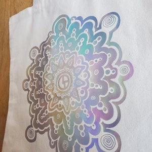 May include: A white canvas tote bag with a large iridescent mandala design. The mandala is a circular pattern with a variety of shapes and swirls.