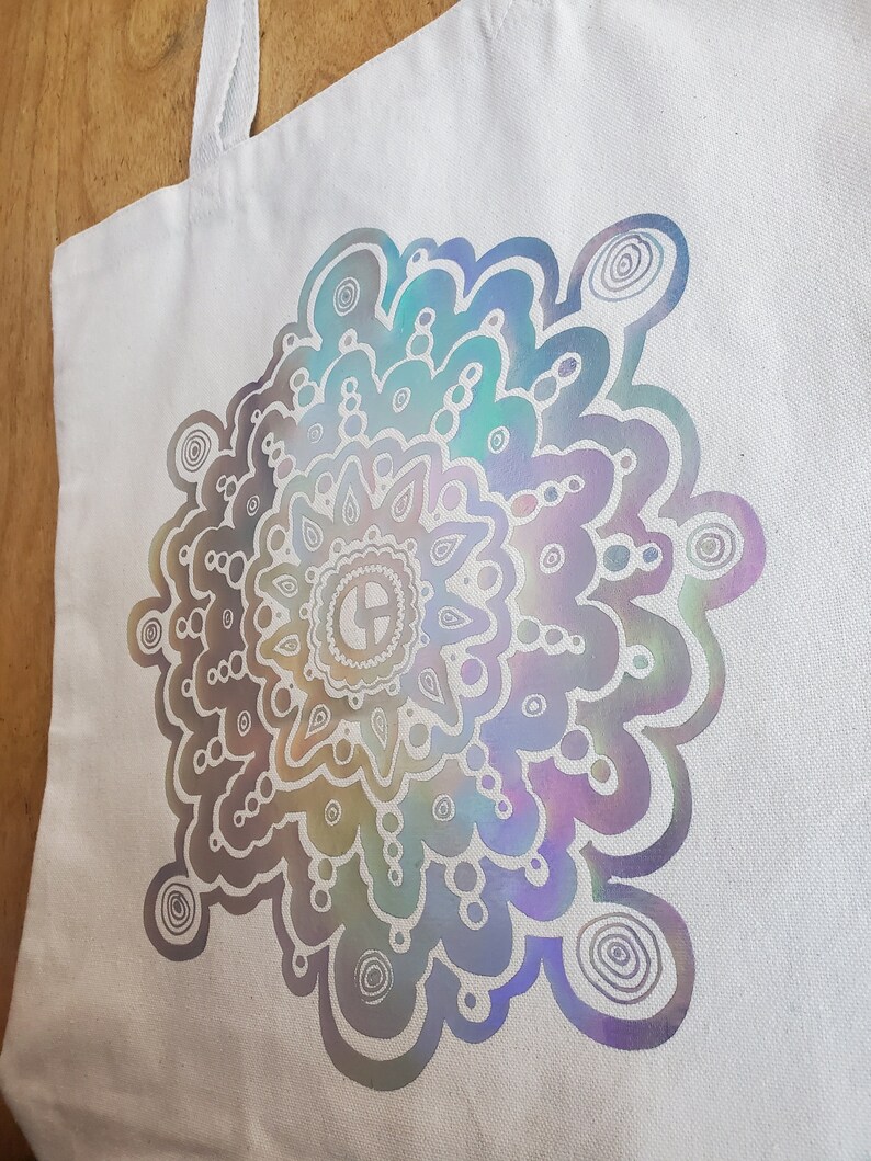 May include: A white canvas tote bag with a large iridescent mandala design. The mandala is a circular pattern with a variety of shapes and swirls.
