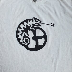 K&ouml;nnte beinhalten: Wei&szlig;es T-Shirt mit einem schwarzen Grafikdruck eines Cham&auml;leons mit langer Zunge, das sich um einen Kreis mit einem stilisierten Buchstaben "E" windet.
