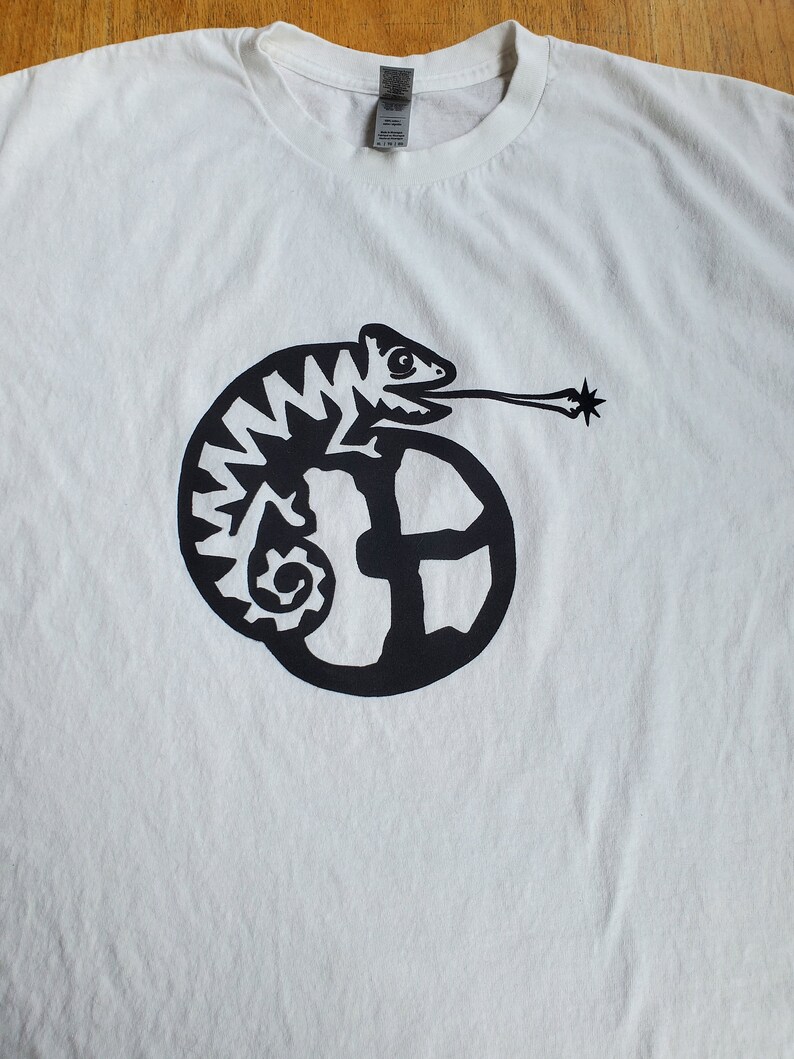 K&ouml;nnte beinhalten: Wei&szlig;es T-Shirt mit einem schwarzen Grafikdruck eines Cham&auml;leons mit langer Zunge, das sich um einen Kreis mit einem stilisierten Buchstaben "E" windet.