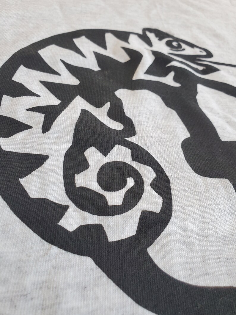 K&ouml;nnte beinhalten: Ein graues T-Shirt mit einem schwarzen und wei&szlig;en Grafikdruck eines Cham&auml;leons. Das Cham&auml;leon ist mit seinem Schwanz im Maul eingerollt.
