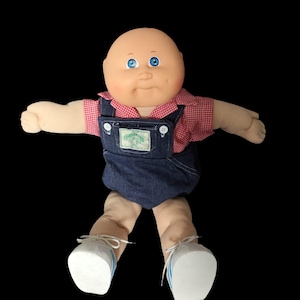 May include: A vintage Cabbage Patch Kid doll with a bald head, blue eyes, and rosy cheeks. The doll is dressed in a red and white checked shirt under denim dungarees. White trainers with blue stripes complete the outfit.