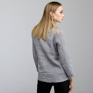 May include: A woman wearing a gray and white striped sweater with long sleeves. She is standing with her back to the camera and looking over her shoulder.