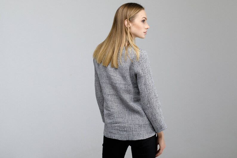 May include: A woman wearing a gray and white striped sweater with long sleeves. She is standing with her back to the camera and looking over her shoulder.
