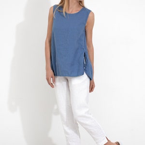 May include: A woman wearing a blue linen sleeveless top with side slits and white linen pants. She is wearing white sandals.