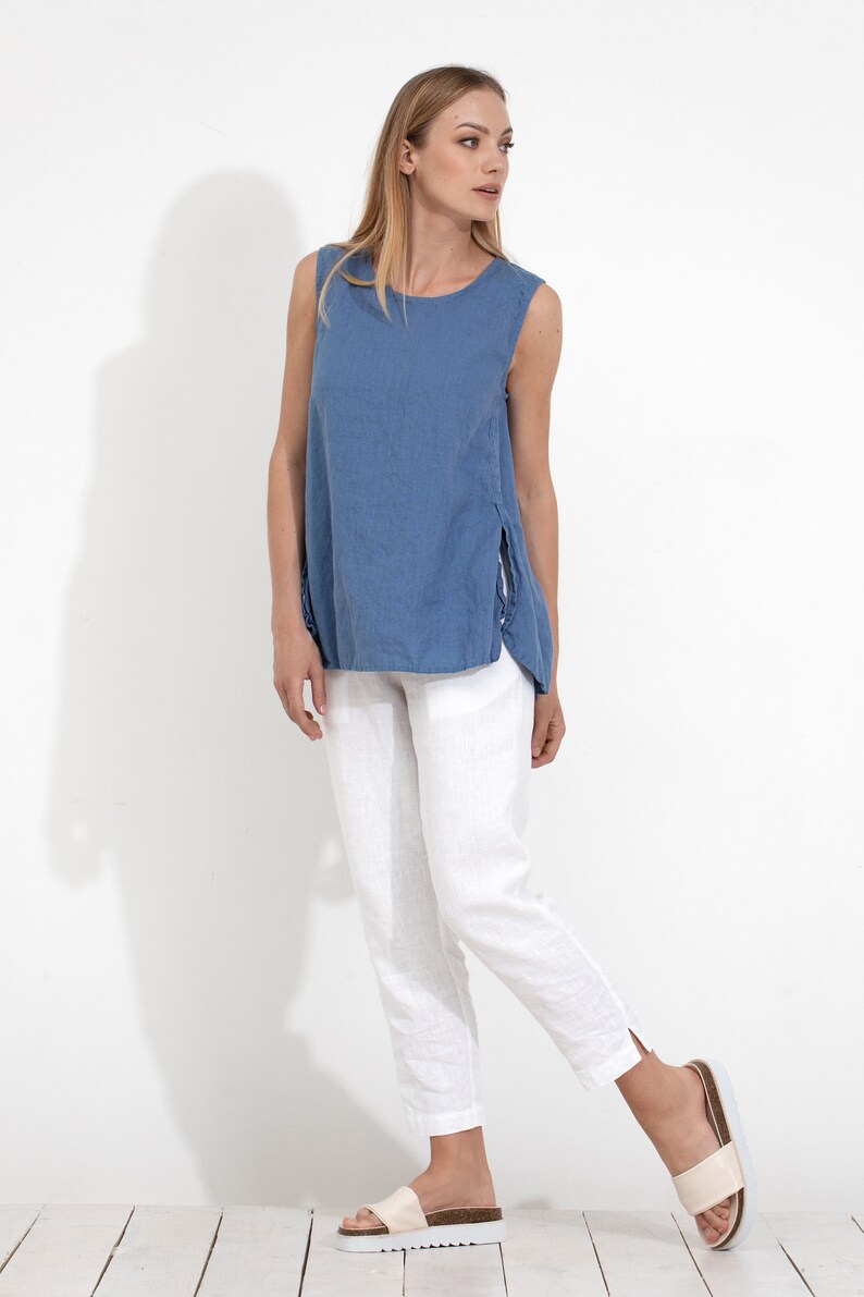 May include: A woman wearing a blue linen sleeveless top with side slits and white linen pants. She is wearing white sandals.