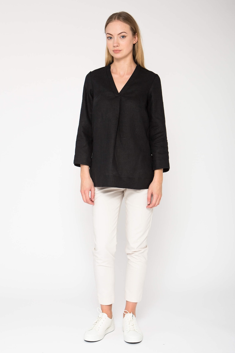 Puede incluir: Una blusa de lino negra con cuello en V y mangas 3/4. La blusa es holgada y tiene una silueta relajada. La modelo lleva la blusa con pantalones blancos y zapatillas blancas.