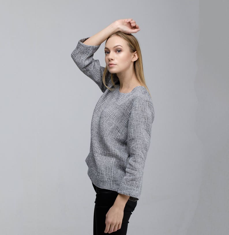 May include: A woman wearing a gray, textured sweater with long sleeves and a round neckline. She is wearing black pants and has her hand on her head.