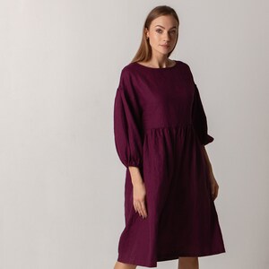 Puede incluir: Un vestido de lino burdeos con cuello barco y mangas 3/4. El vestido tiene una cintura fruncida y una falda ligeramente acampanada.