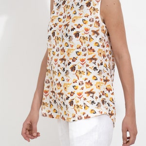 May include: A sleeveless white top with a floral print featuring yellow, orange, and brown flowers. The top has a round neckline and a relaxed fit.