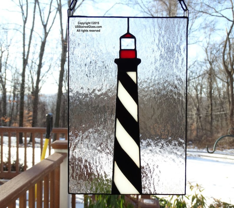 May include: A stained glass window depicting a black and white striped lighthouse with a red roof against a textured glass background.