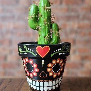 May include: A green cactus with brown spines in a black Day of the Dead themed plant pot. The pot has a skull design with red, orange, and white floral and heart accents. The background is a brick wall.