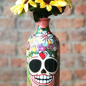 May include: A pink glass bottle with a painted design of a smiling skull wearing a floral crown and a turquoise dress. The bottle is filled with yellow sunflowers.