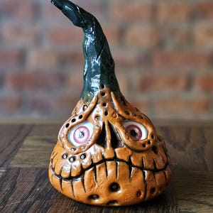 Może przedstawiać: Pomarańczowa ceramiczna dekoracja na Halloween w kształcie dyni z groźną twarzą i wypukłymi oczami. Dynia ma ciemnozieloną łodygę i jest ustawiona na tle ceglanej ściany.