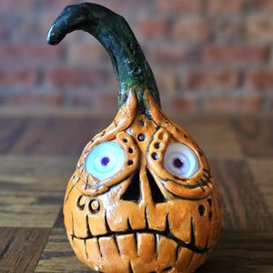 Pode incluir: Uma abóbora de cerâmica laranja artesanal com um rosto esculpido e um caule verde curvo. A abóbora tem olhos grandes e expressivos com íris azuis e roxas e um sorriso largo. Uma decoração única de Halloween.
