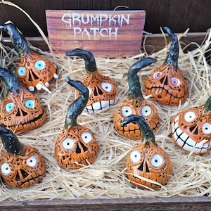 Puede incluir: Una caja de madera llena de paja y varias figuras de cerámica naranja con forma de calabaza con caras talladas y tallos verdes curvados. El letrero en el fondo dice "GRUMPKIN PATCH". Las calabazas tienen dientes blancos y ojos azules o blancos.