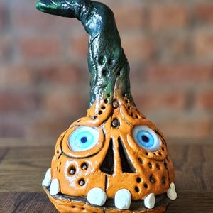 Może przedstawiać: Kapryśna pomarańczowa ceramiczna rzeźba w kształcie dyni z zieloną, zakrzywioną łodygą. Posiada niebieskie oczy, uśmiech i dekoracyjne otwory. Unikalny element dekoracyjny na Halloween lub jesień.