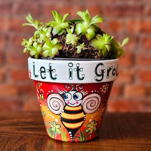 May include: A red and white ceramic planter with a bee design and the words "Let it Grow" painted on the side. The planter is filled with green succulents.
