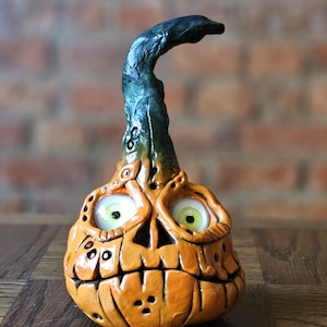 Puede incluir: Una escultura artesanal con forma de calabaza naranja con una cara espeluznante y un tallo verde. La calabaza tiene ojos grandes y expresivos con iris verdes y una boca tallada. La escultura es probablemente de cerámica o arcilla.
