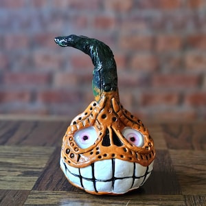 May include: A handcrafted Halloween gourd sculpture with an orange body, a green stem, and a grinning face with white teeth and pink eyes. The gourd has carved details and a textured surface, ideal for seasonal decor.