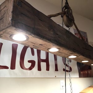 May include: A rustic wooden beam light fixture with four integrated lights. The beam is dark brown with a natural wood grain and is suspended from the ceiling by a rope and pulley system.