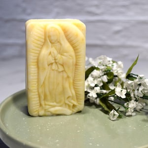 May include: A rectangular, light yellow soap bar with a carved image of a religious figure. The soap sits on a light green plate, with white flowers and green leaves to the side. The soap's design is detailed and the overall aesthetic is serene.