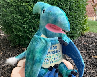 May include: A plush pterodactyl toy in shades of blue and green, with a pink mouth and black claws. The toy is embroidered with the name "Alan Cleveland", birthdate, time, weight, and length of 20 inches. It sits on a wooden slice.