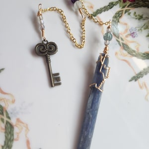 Puede incluir: Un collar hecho a mano con un cristal de cianita azul envuelto en alambre dorado, conectado por una cadena dorada a un dije de llave de bronce. La cadena también tiene cuentas de varios colores. El collar se exhibe en un plato con estampado floral.