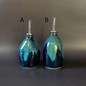 May include: Two ceramic oil bottles with stainless steel spouts. The bottles are teardrop-shaped, with a dark blue base and a turquoise and blue glaze that drips down the sides. The letters "A" and "B" are printed above the bottles.