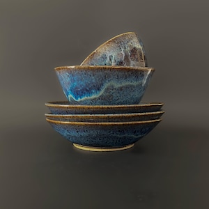 May include: A stack of three ceramic bowls featuring a reactive glaze with deep blue, teal, and brown tones. The bowls have a speckled texture and a dark brown rim, with one bowl positioned at an angle on top of the others in the stack.