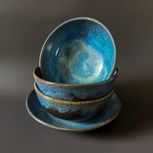 May include: A stack of three ceramic bowls and a plate. The bowls and plate are glazed in shades of blue and brown, with a glossy finish. The bowls are stacked on top of each other, with the plate at the bottom.