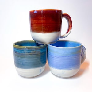 May include: Three ceramic mugs with different colored glazes. The top mug is red with white glaze at the bottom. The bottom two mugs are blue with white glaze at the bottom.