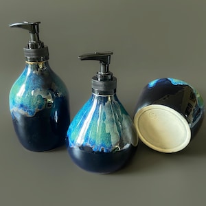 May include: Three ceramic soap dispensers with a glossy finish. The dispensers are a dark blue at the base, transitioning to turquoise and blue, and topped with a black pump. One dispenser is lying on its side, revealing a white interior.
