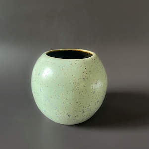 May include: A round, speckled vase with a light green exterior and a dark brown interior. The ceramic vase has a smooth, spherical shape and a wide opening. The surface is dotted with small, dark specks.