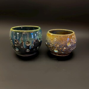 May include: Two handmade ceramic cups with a unique textured design. One cup is a deep blue with green and black accents, while the other is a mix of brown, gold, and blue. Both have a series of small, circular openings. The cups are on a black background.