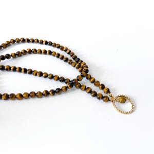 以下が含まれることがあります： 茶色と金色の小さな丸いタイガーアイの石を使ったビーズネックレス。 ネックレスには、楕円形のタイガーアイの石と装飾的な金色のフレームが付いた、金色のペンダントが付いています。