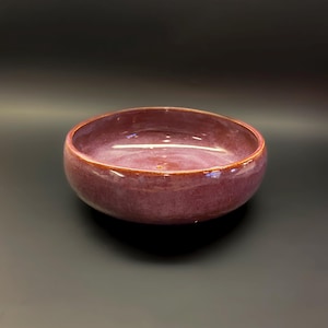 May include: A handmade ceramic bowl with a deep, rounded shape. The bowl features a rich, mottled plum color with a glossy finish. The rim has a contrasting reddish-brown hue, adding visual interest. This decorative bowl is suitable for serving or display.
