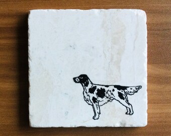 May include: Square white marble coaster featuring a black and white illustration of a dog. The dog is standing, with a spotted coat and a detailed outline. The coaster is set against a wooden surface.