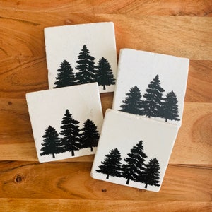 May include: Set of four white square coasters with black silhouette images of three pine trees on each coaster.