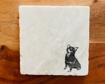 Op de afbeelding: Vierkante, gebroken witte stenen onderzetter met een zwart-wit illustratie van een Chihuahua hond. De hond zit en kijkt omhoog. De onderzetter heeft een getextureerd oppervlak en ligt op een houten achtergrond.