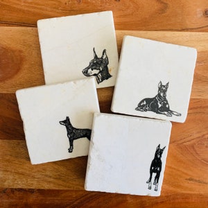 May include: Four white square coasters with black and white images of Doberman Pinschers. The coasters are made of natural stone and have a rustic look.