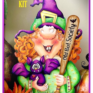 May include: A wooden craft kit featuring a witch with orange hair, a green shirt with stars, a purple hat, and a purple bat. She is holding a wooden baseball bat with the words "Old Bat Society" written on it. The kit is titled "Old Bat Society KIT".